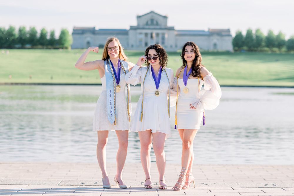 Three graduates in white dresses stand together by a lake holding medals, with a stately building in the background.