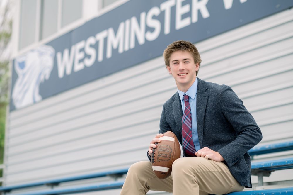 Student athlete in a suit and tie sits on bleachers holding a football at Westminster with building signage visible.
