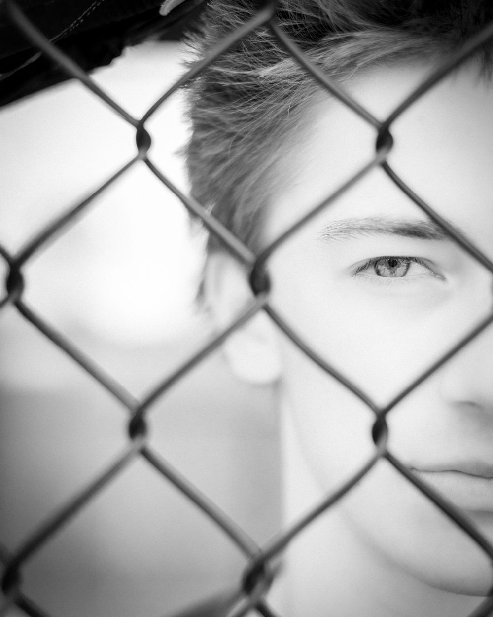 Artistic black and white portrait through chain link fence creates moody composition with dramatic lighting and shadow.