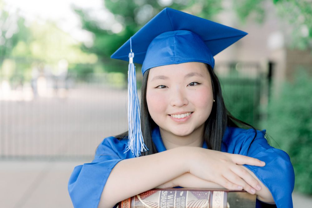 Graduate wearing traditional blue cap and gown smiles while posing with a graduation diploma on campus.