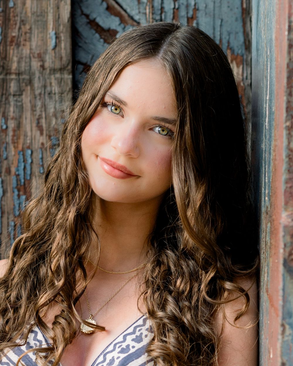 Woman with long dark curly hair poses near rustic wooden wall wearing a patterned top.