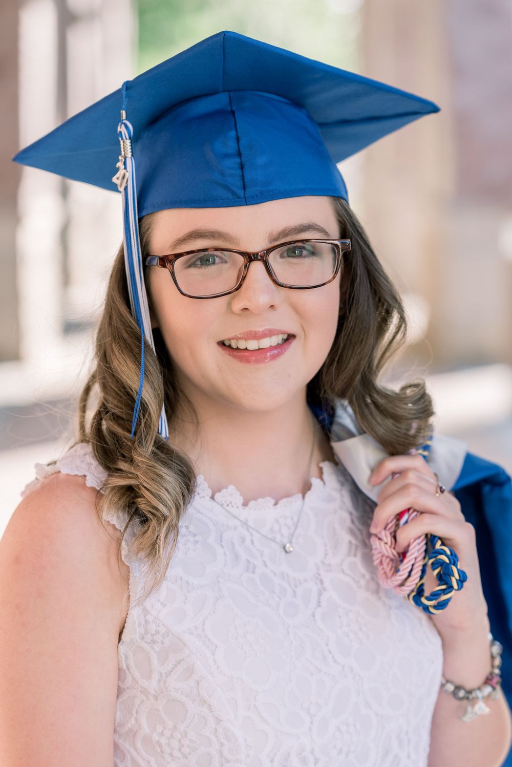 Graduation portrait showing a graduate in blue cap and white lace dress wearing glasses with hair styled in loose curls.