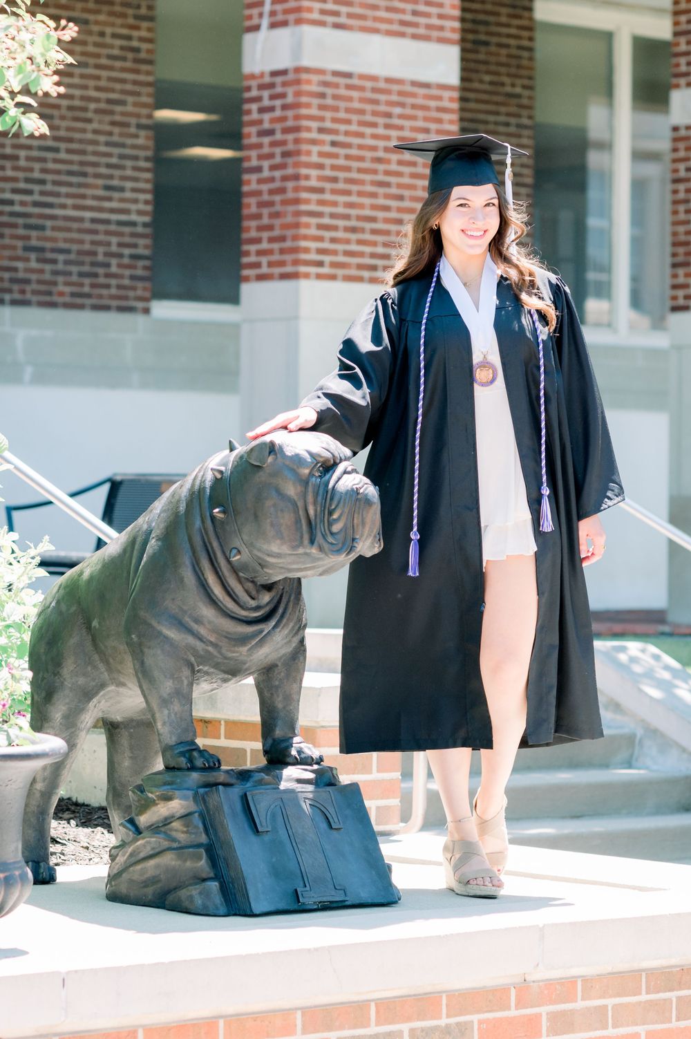 Graduate in cap and gown poses next to bronze bulldog statue with a backpack on brick steps of university building.