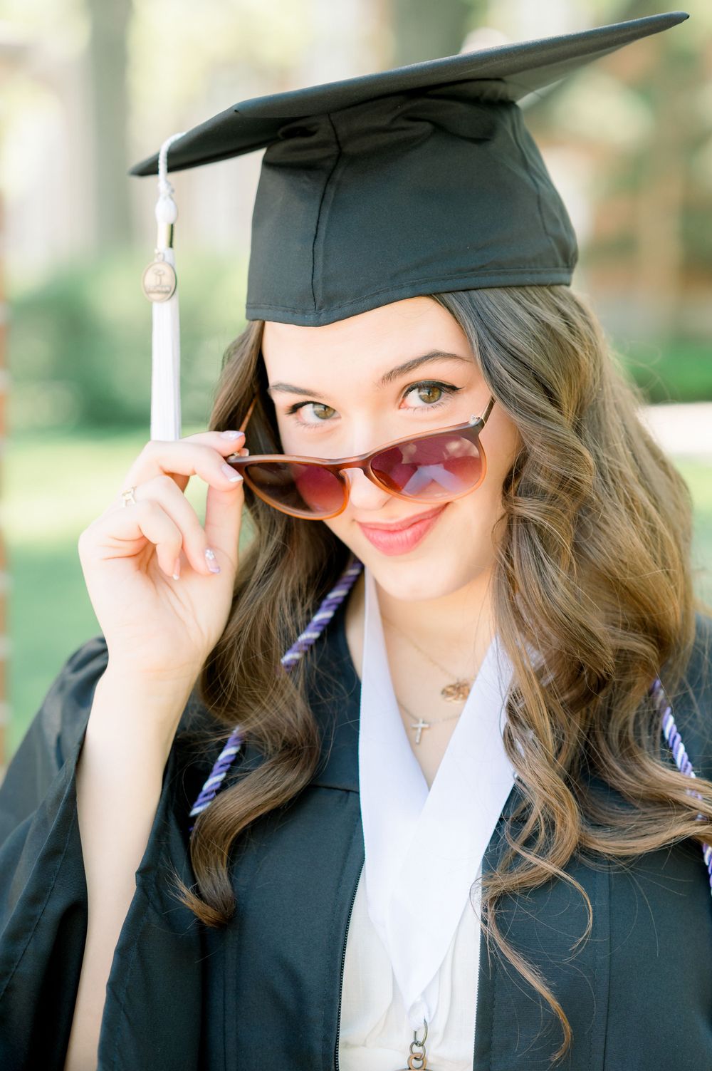 A graduate in cap and gown peers over sunglasses with a playful expression during a graduation photoshoot.