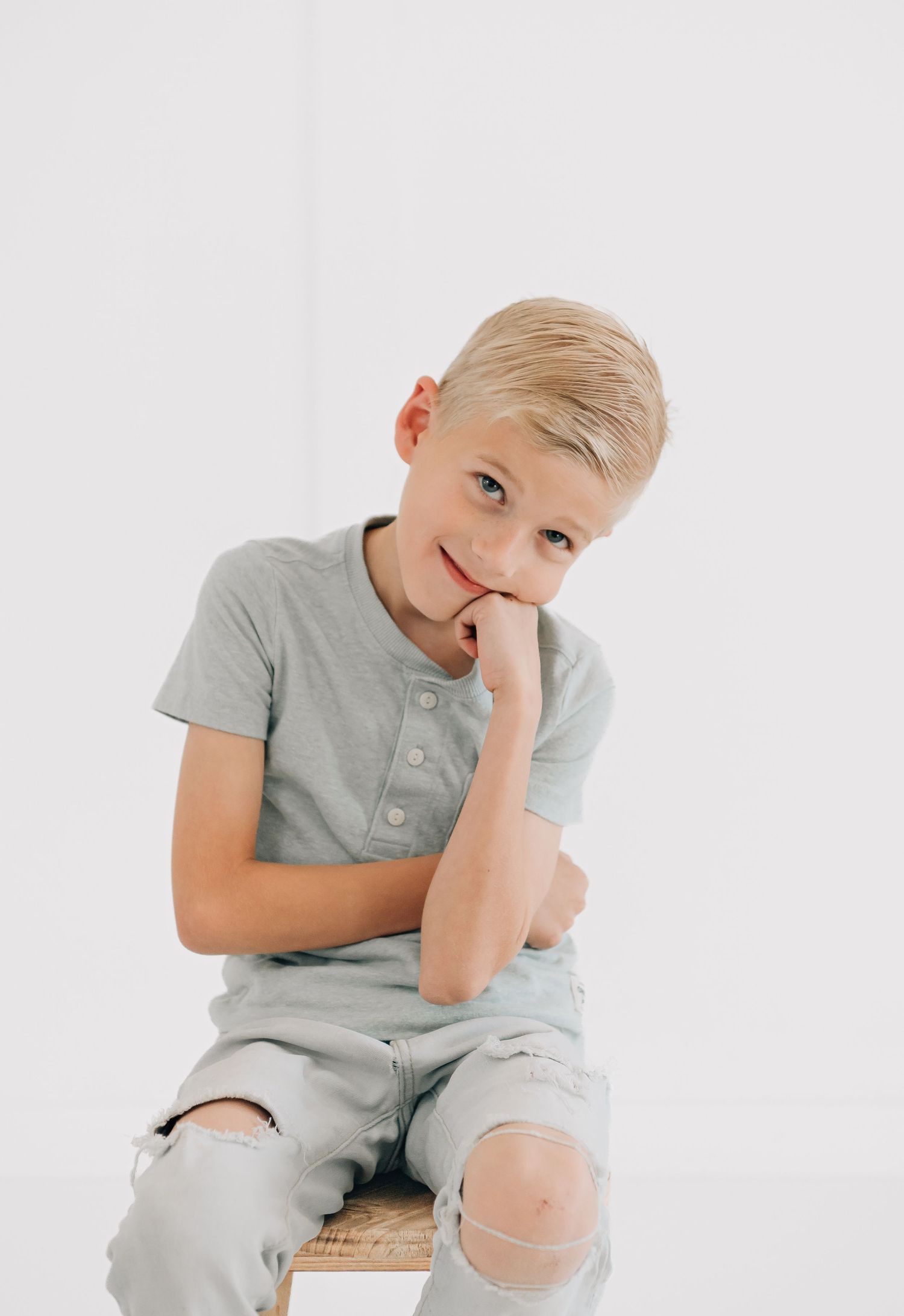 Blonde boy in gray henley shirt and ripped jeans sits on wooden stool, resting chin on hand, smiling.