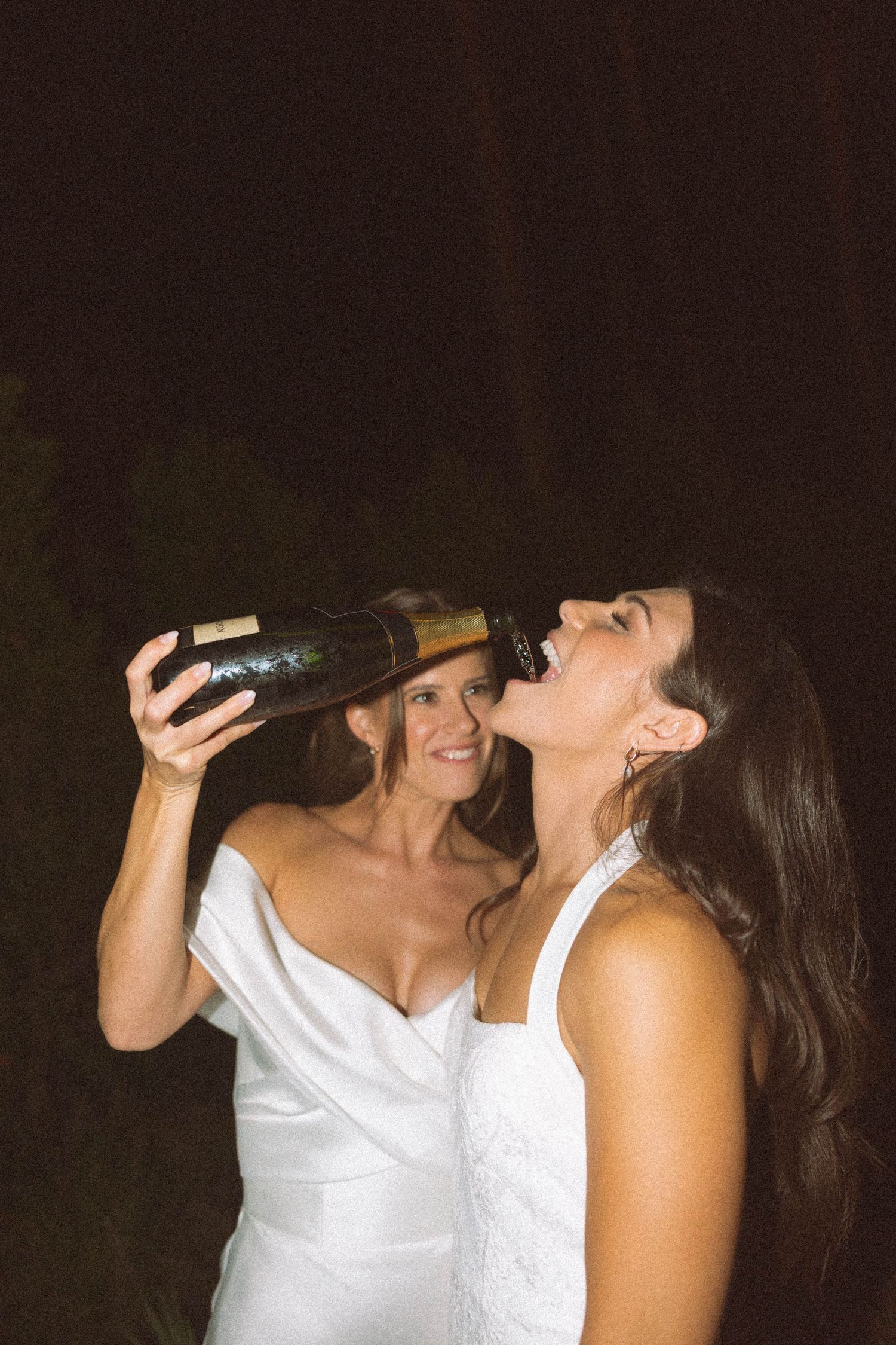 Two people in white dresses sharing a celebratory drink from champagne bottles at a festive event.