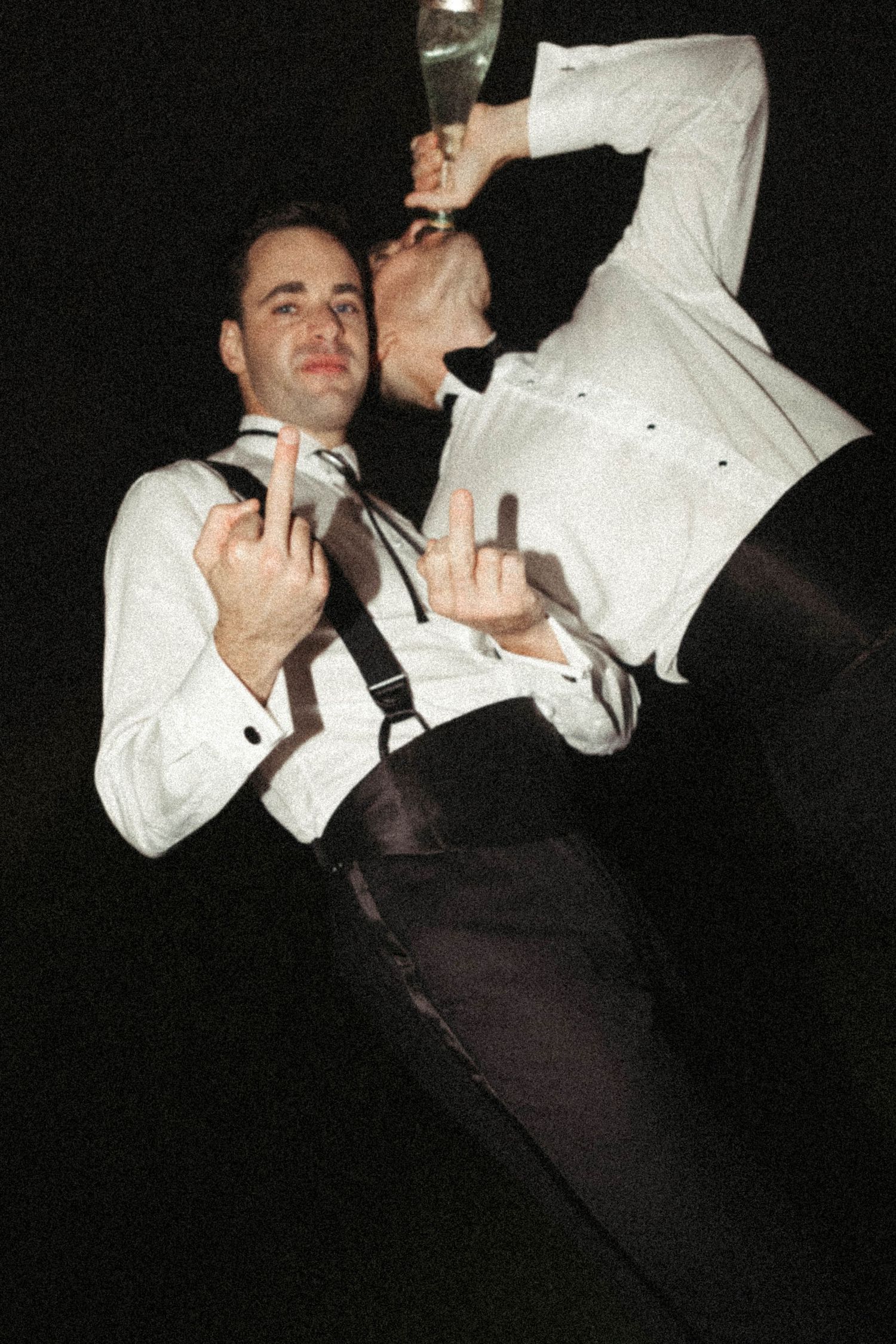 Two men in white dress shirts making rude gestures at the camera during what appears to be a party or event.
