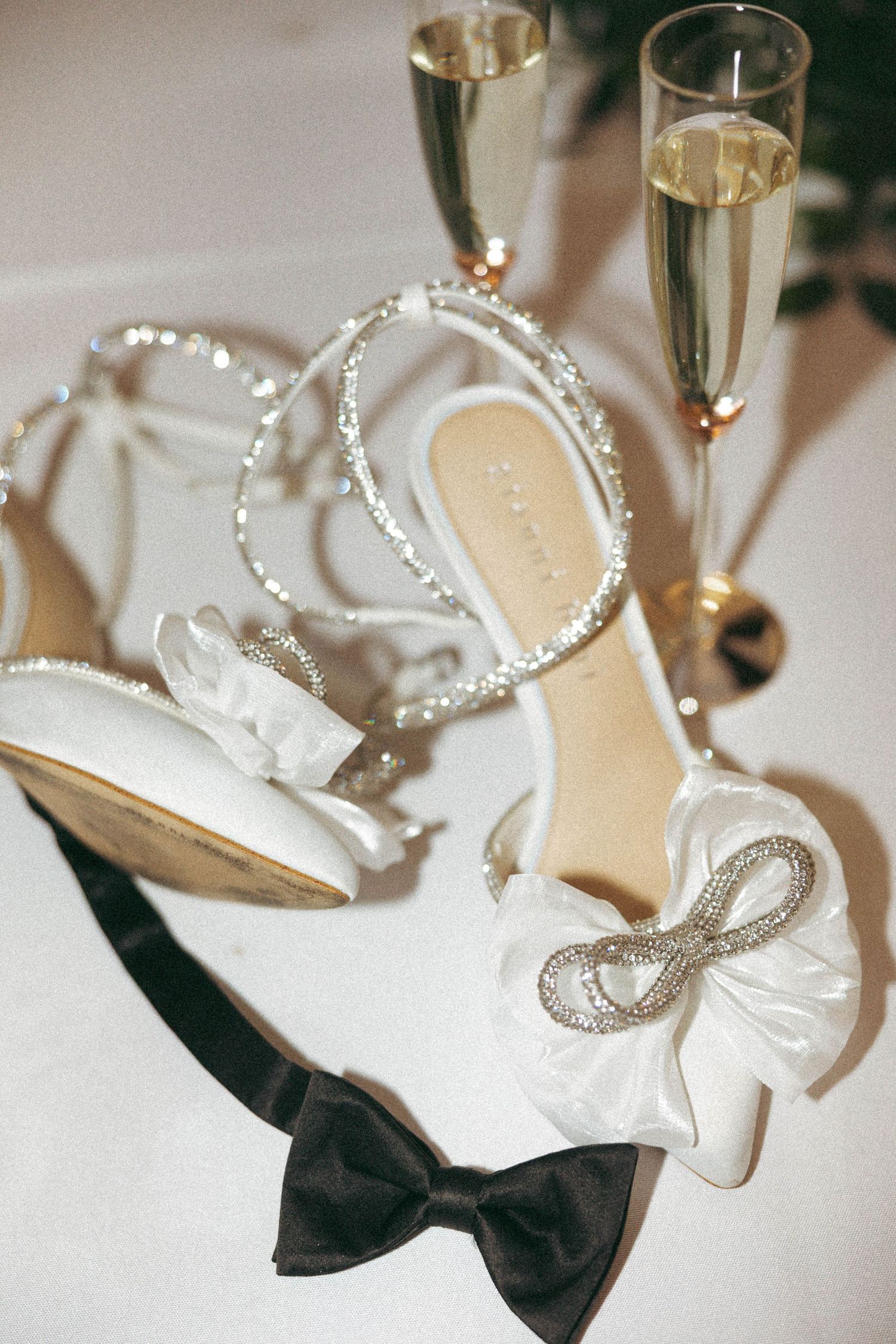 White bridal shoes with pearl straps and black bow details sit elegantly next to champagne glasses at a wedding celebration.