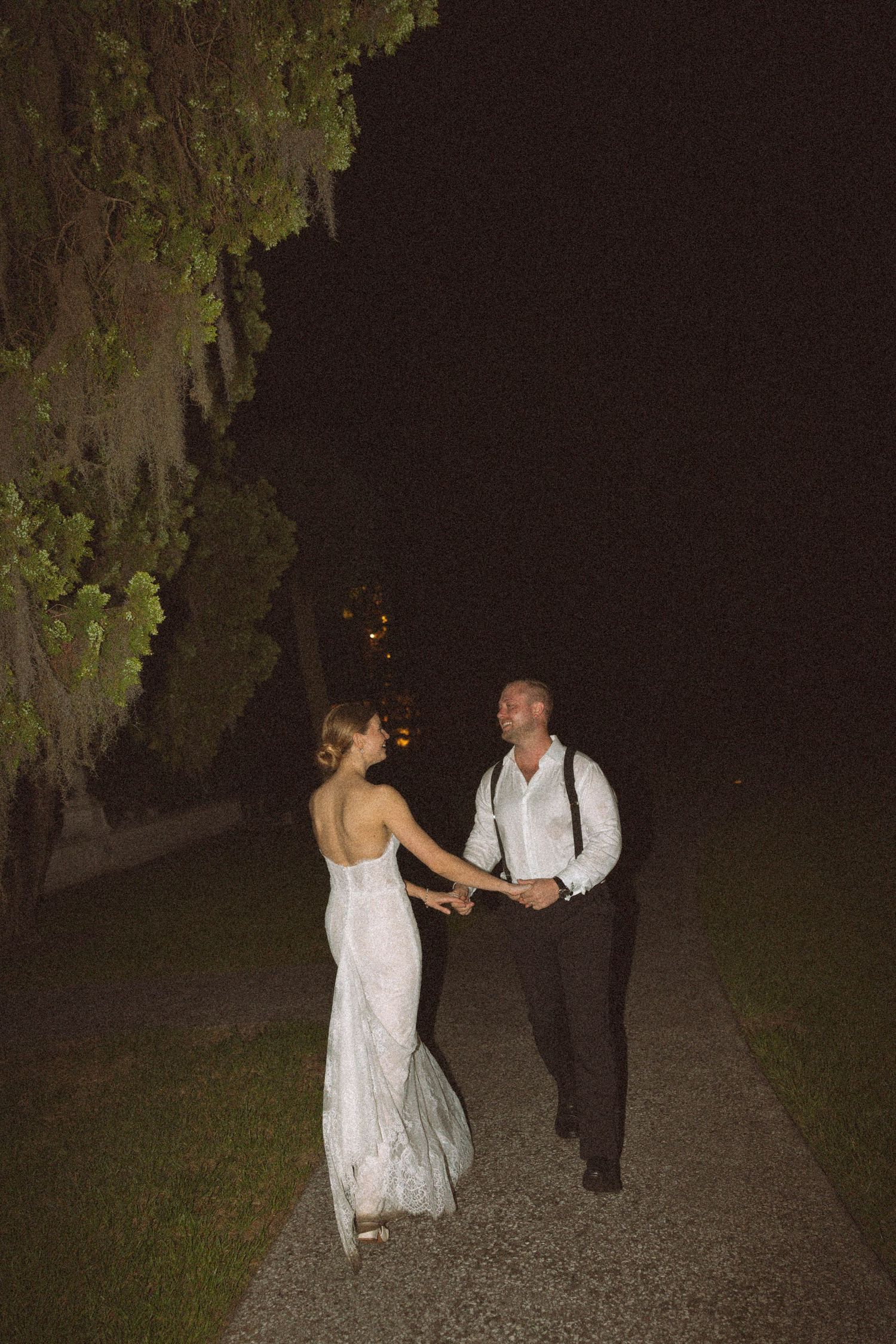 A couple in wedding attire walk together on a dark gravel path under trees at night.