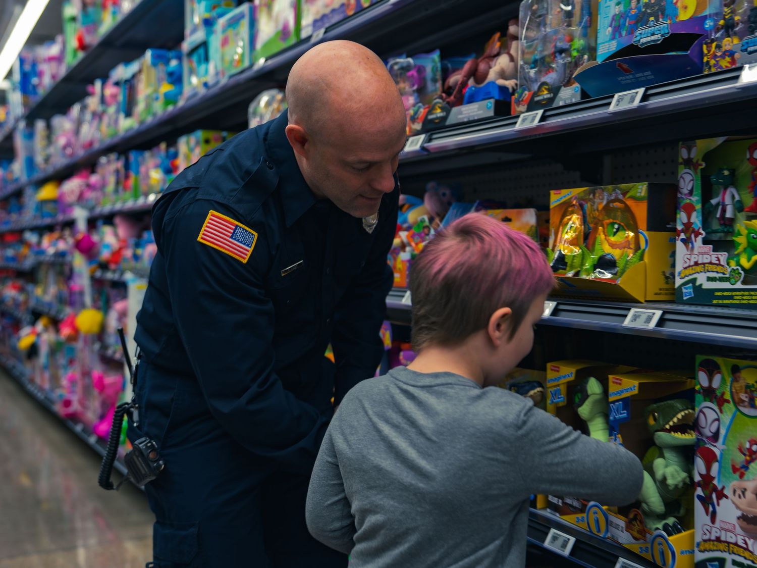 A firefighter in a navy uniform leans down beside a young child with pink hair who is reaching for a toy on the shelf at the event.