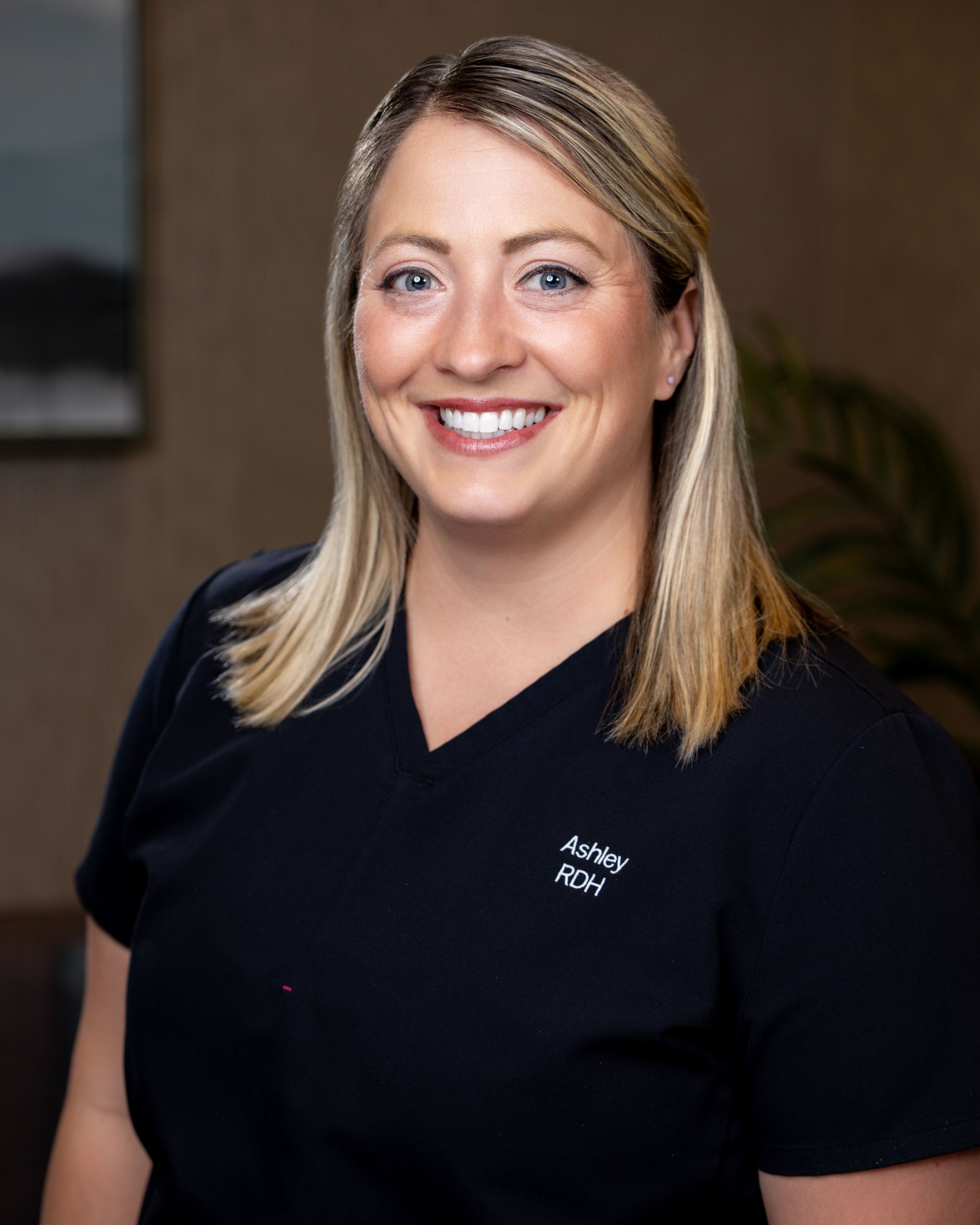 Ashley, RDH dental hygienist, smiling professionally in dark navy scrubs against a warm studio background.