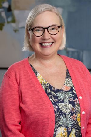 Smiling middle-aged woman with glasses and blonde hair wearing a coral cardigan and floral top.