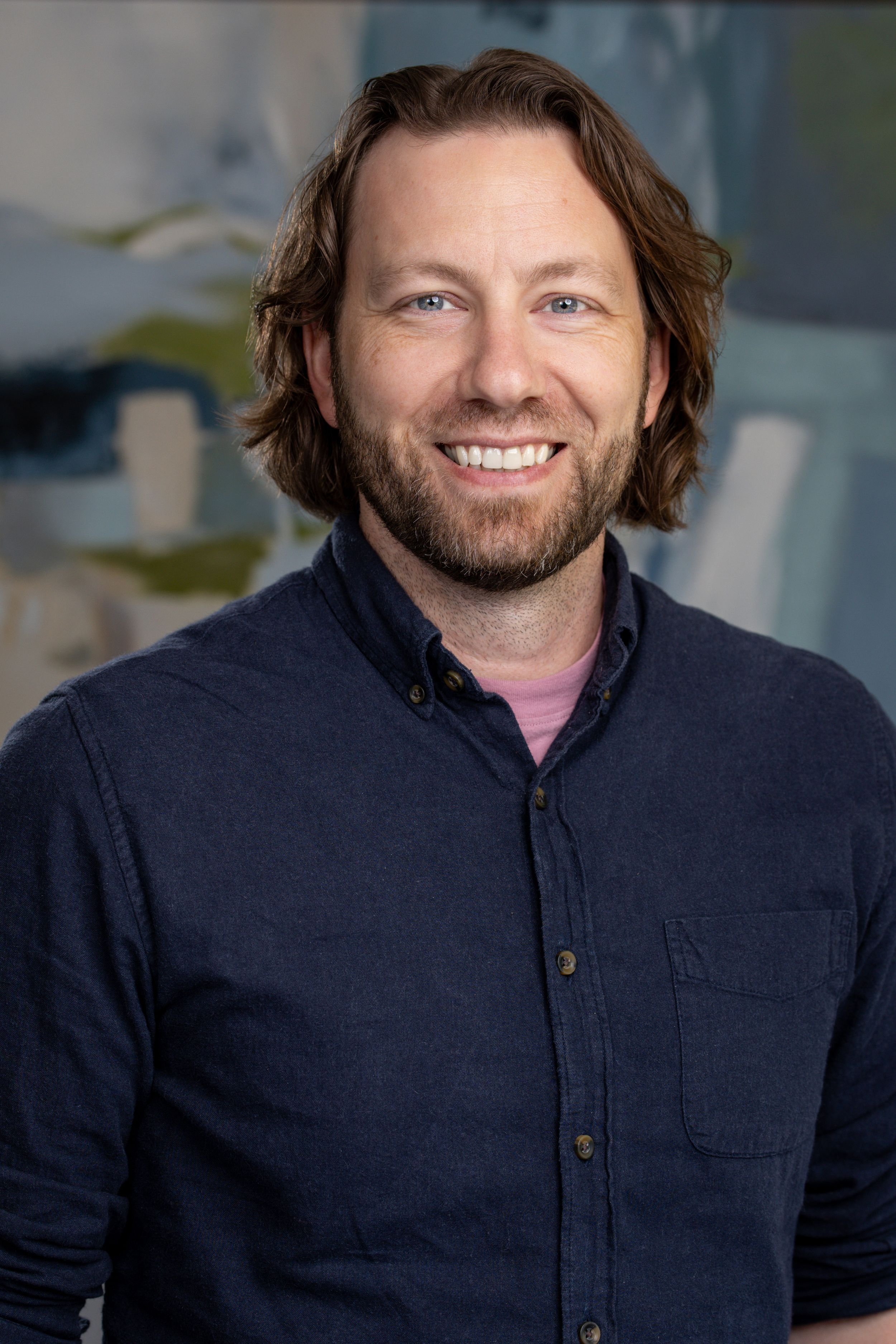 Smiling man with brown hair and beard wearing a navy shirt, posed in front of abstract artwork.