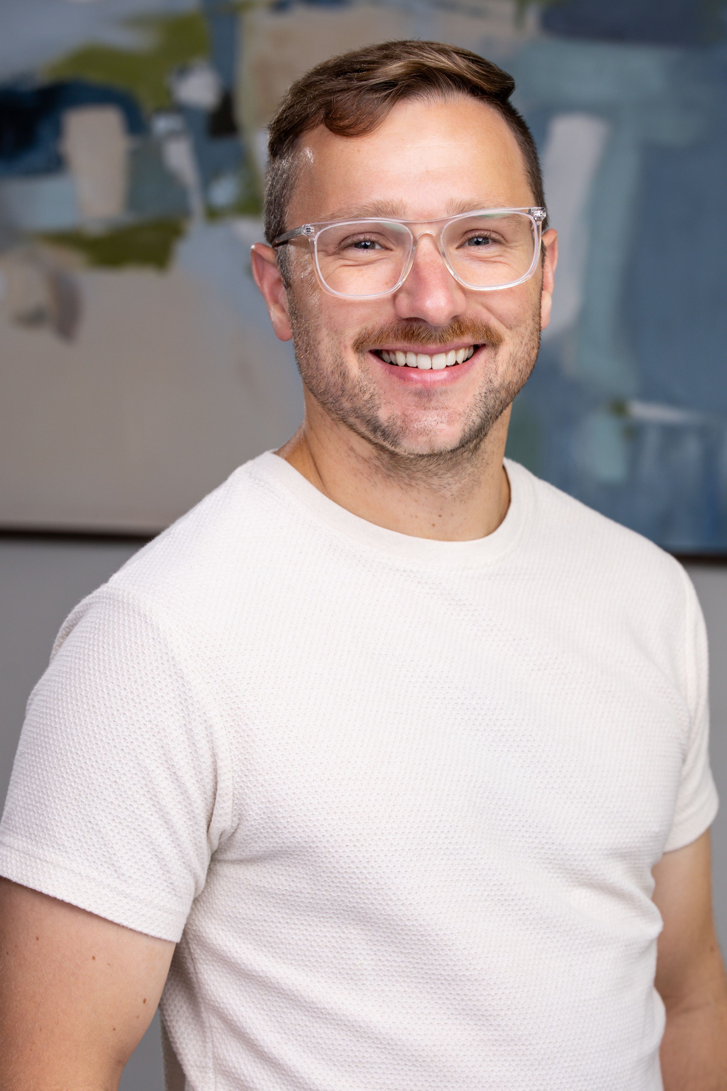 Smiling man with clear glasses and short hair wearing a white t-shirt, posing in front of abstract blue artwork.