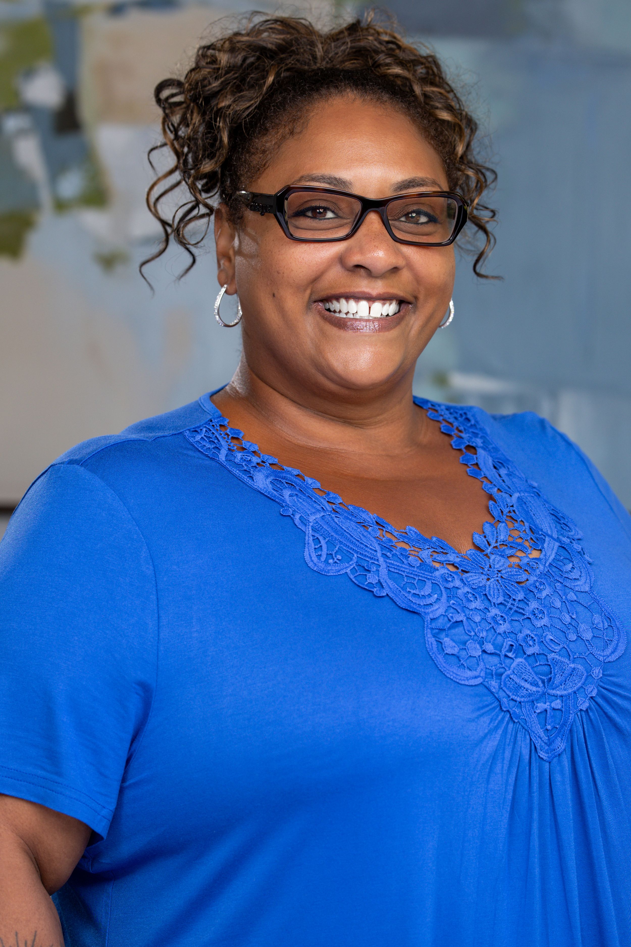 Smiling woman with curly hair and glasses wearing a blue lace v-neck top poses for a professional headshot photo.