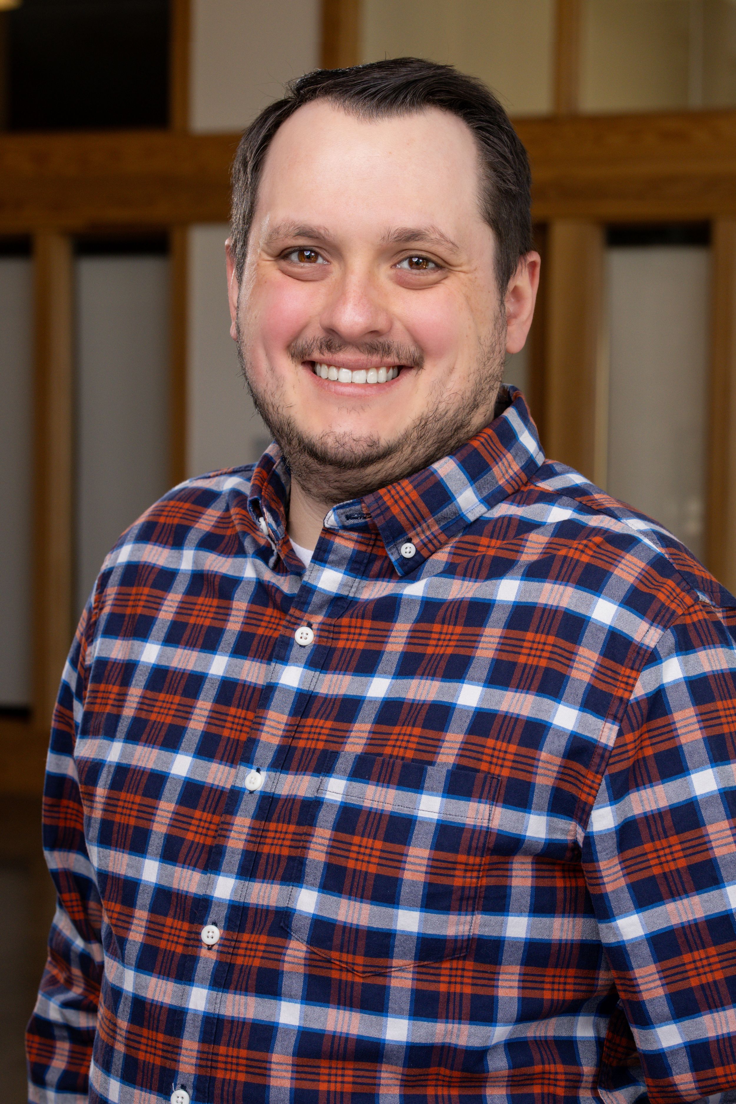 Smiling man in blue and red plaid shirt poses for professional headshot photo indoors.