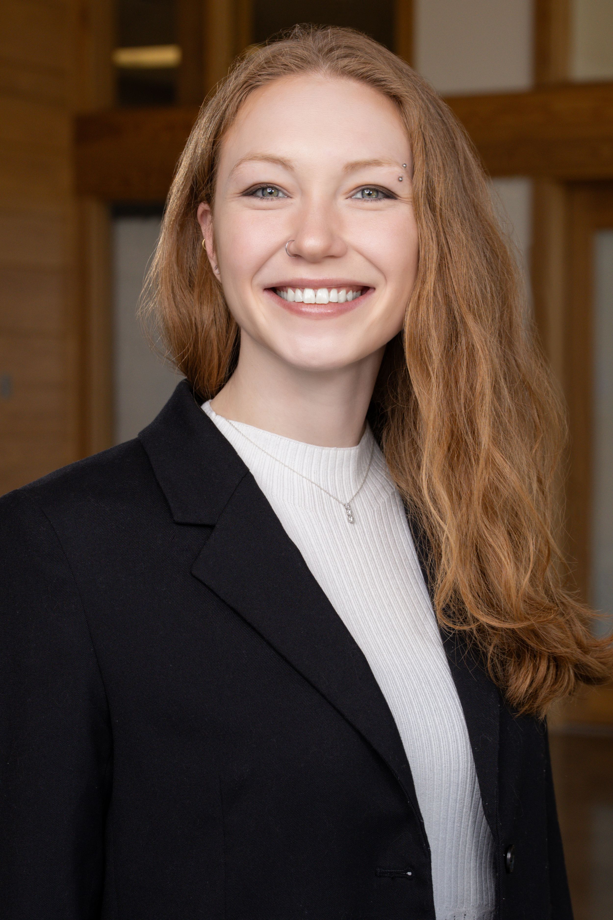 Smiling young professional woman with red hair wearing a black blazer and white turtleneck in a corporate headshot.