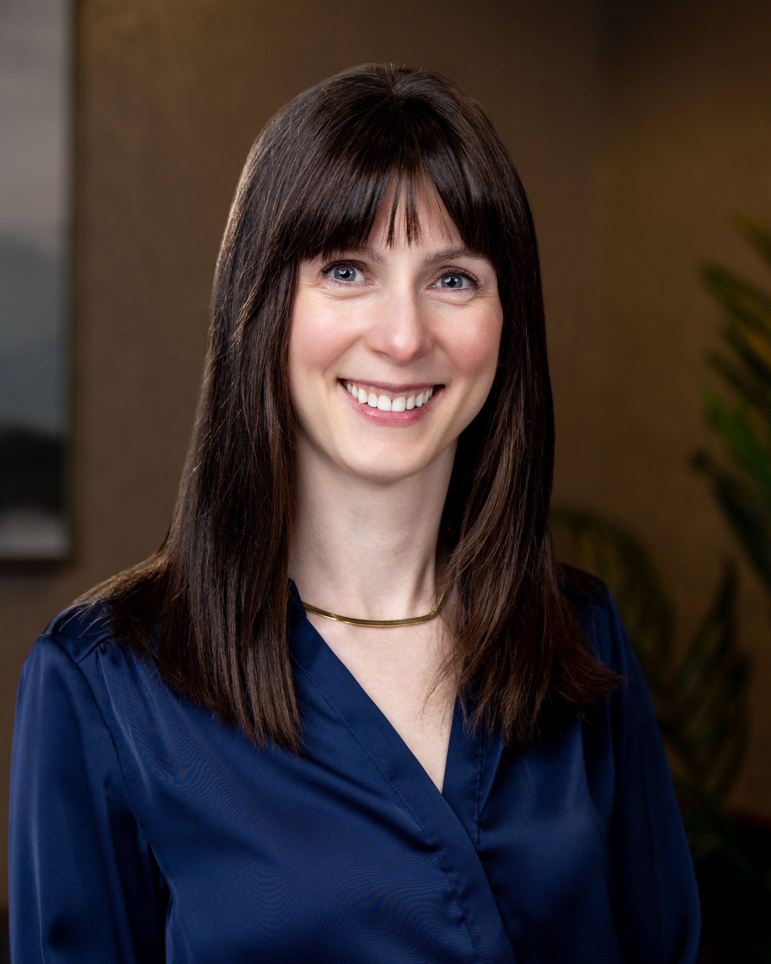 Professional headshot of a smiling woman with dark hair, wearing a navy blue top and gold necklace.