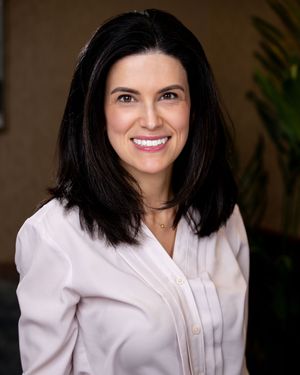 Professional headshot of a smiling woman with dark hair wearing a white blouse in a corporate setting.