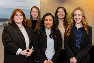 Five professional women in business attire smiling together for a group portrait in an office setting.