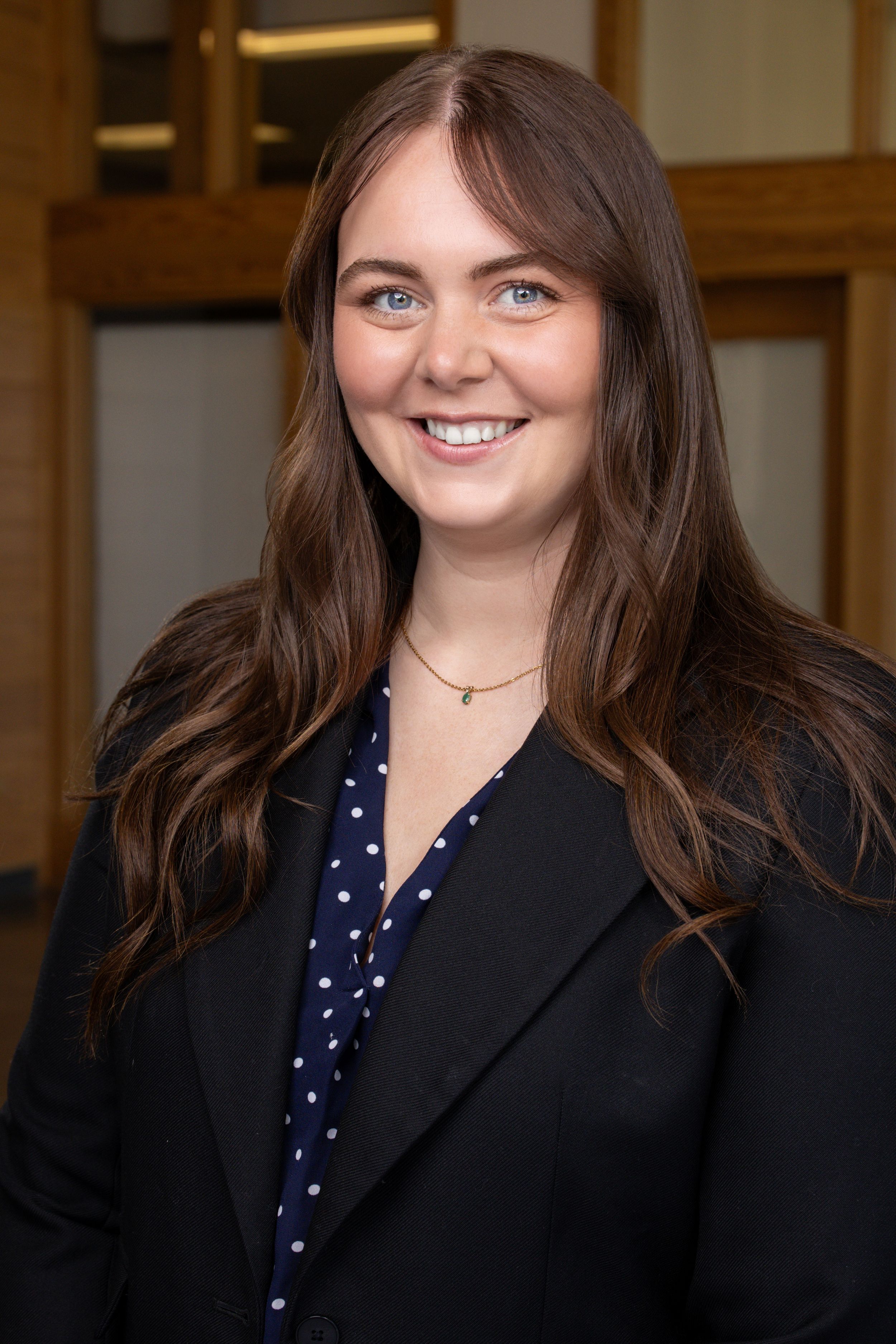 Young professional woman with long brown hair smiling in a black blazer and polka dot blouse.