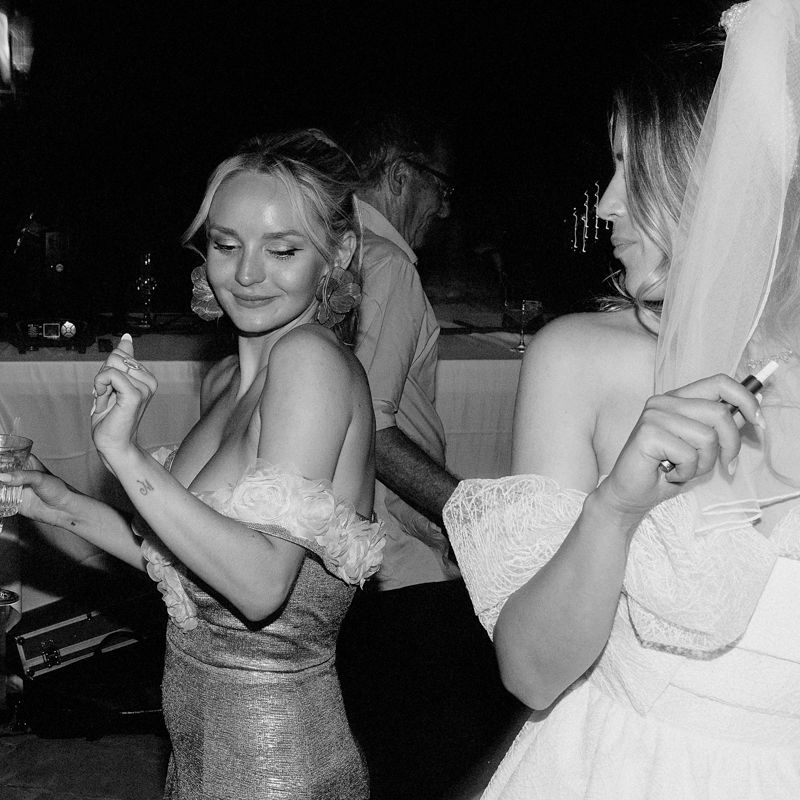 Two women dancing joyfully at a wedding reception, one in a bridal gown, captured in elegant black and white photography.