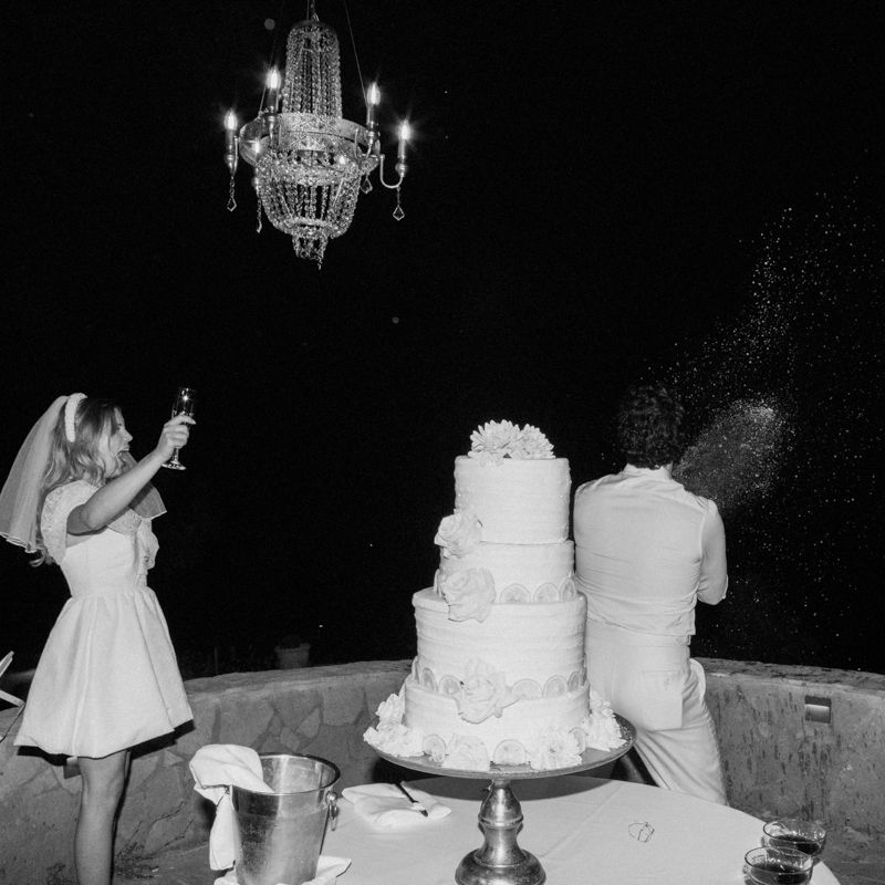 Black and white photo of bride and groom cutting a tiered wedding cake under a chandelier.