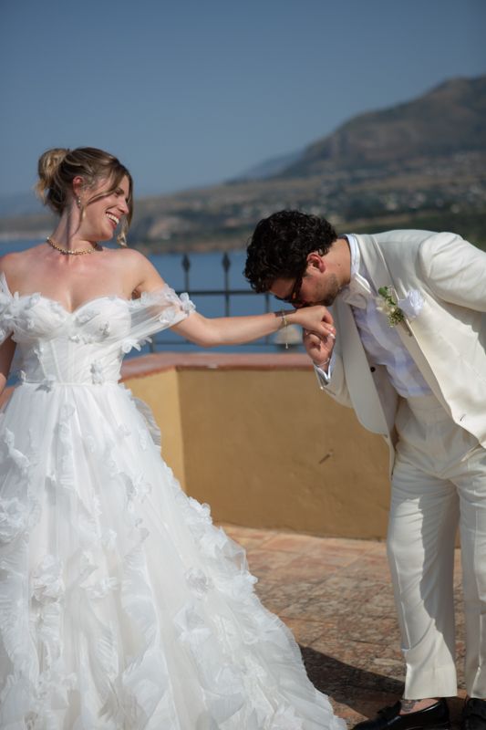 Groom in ivory suit kisses bride's hand as she smiles in a ruffled white gown, with a scenic coastal backdrop.