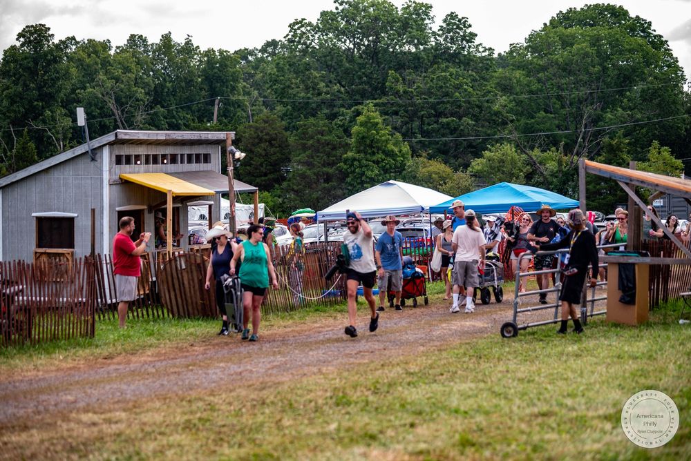 Crowd of people walking through a rustic outdoor farm festival with tents and wooden fences on a cloudy day.