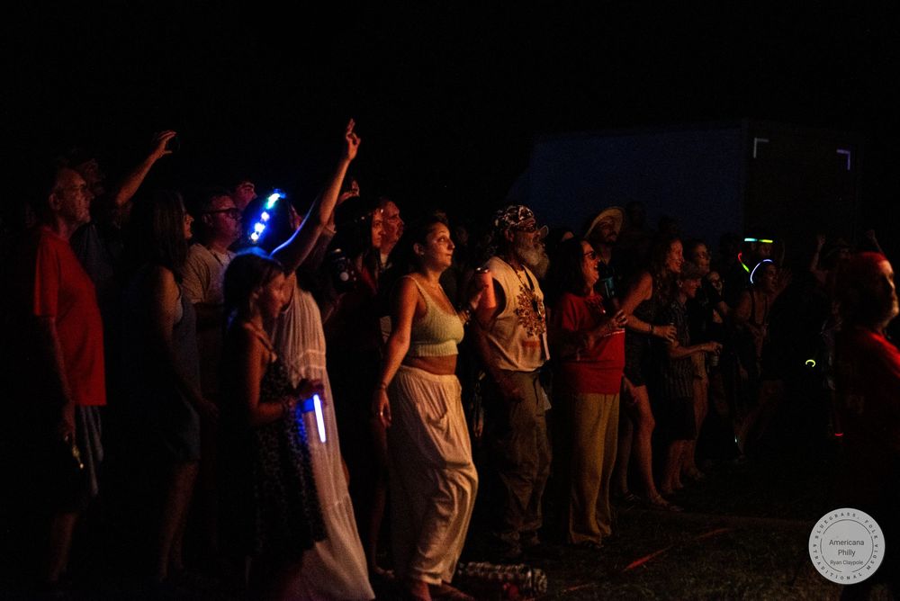 Crowd of festival-goers dancing and cheering at an outdoor night event with colorful glow sticks.