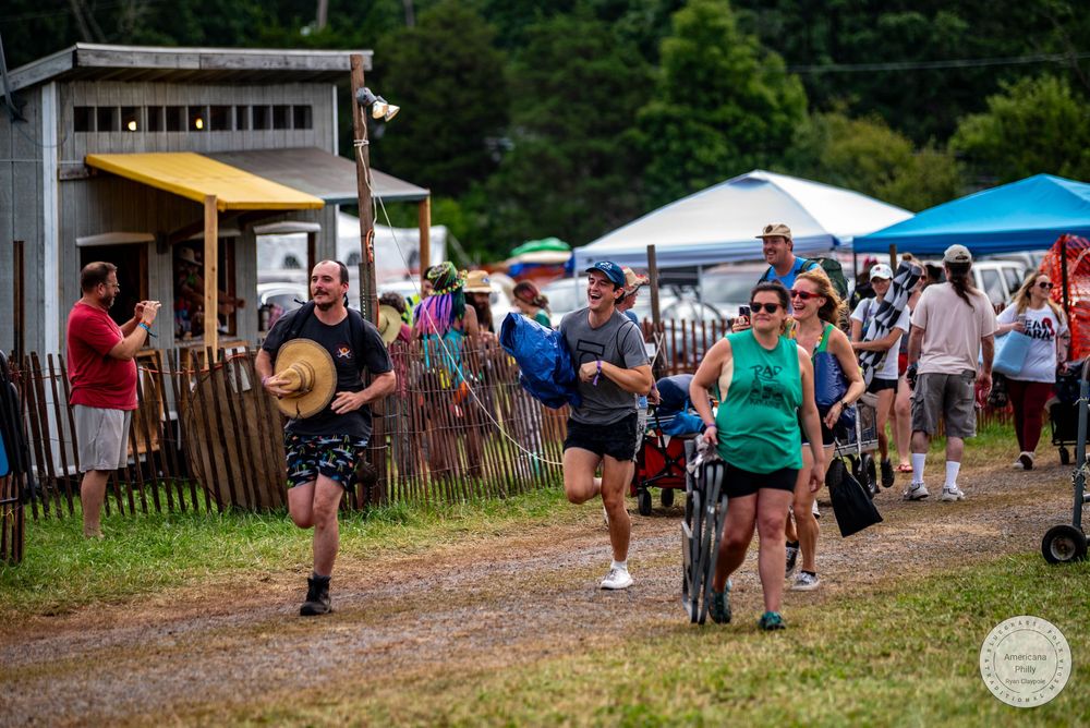 Crowd of people walking at an outdoor festival on a gravel path, with tents and green trees in the background.