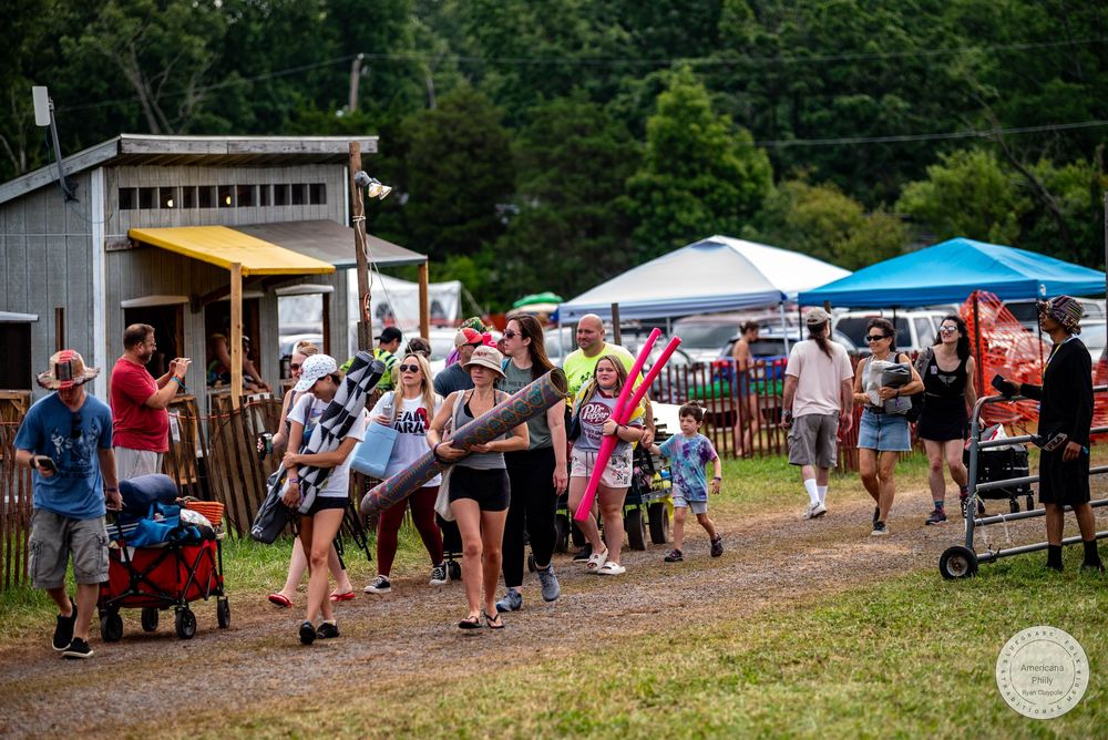 Crowd of people walking at an outdoor summer festival with tents and green grass in a rural setting.