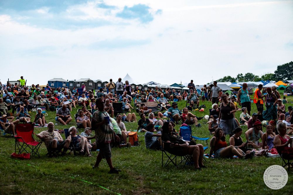 Large crowd enjoying an outdoor music festival on a grassy hill under cloudy skies with colorful chairs and tents.