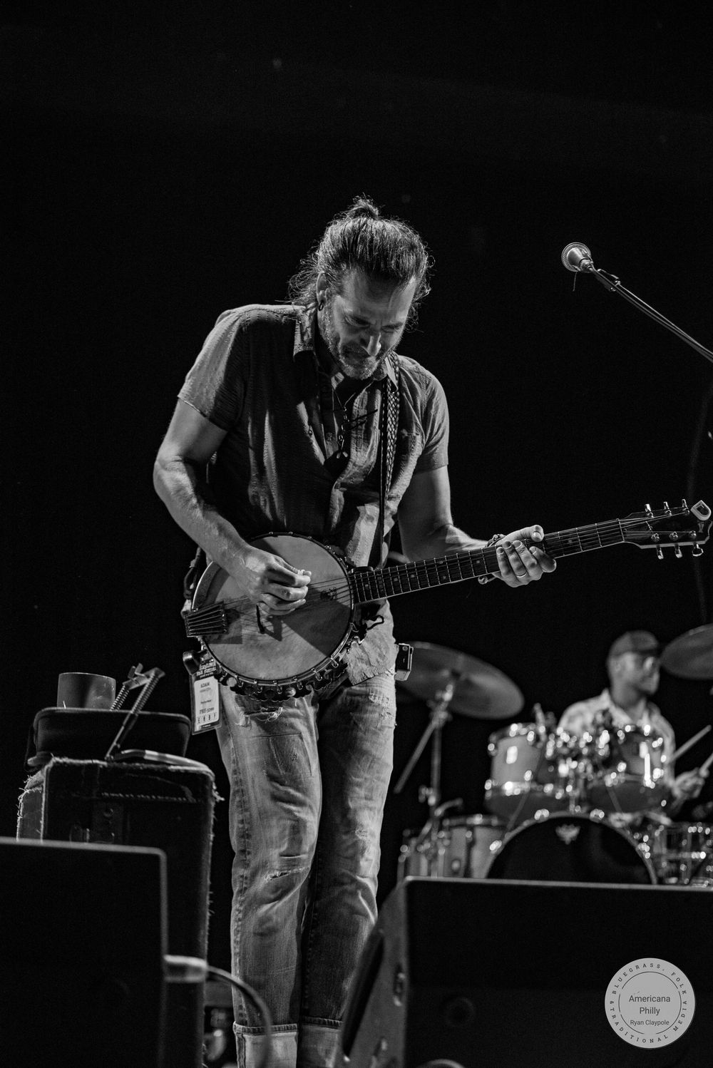 Black and white photo of a long-haired male musician playing a banjo guitar on stage with a drummer visible behind him.