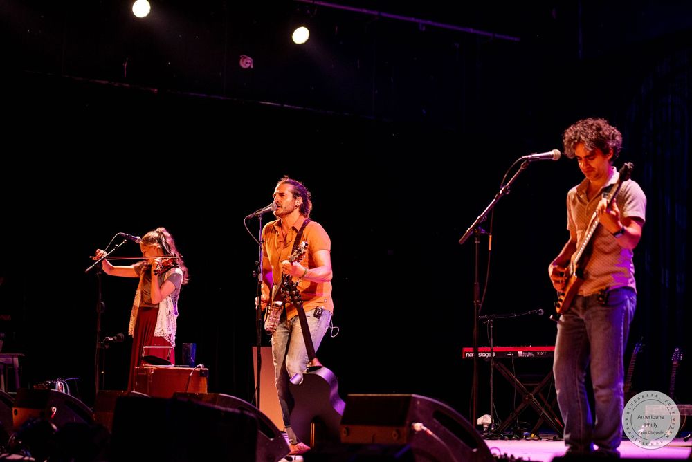 Three musicians performing on stage with guitars and microphone stands under colorful stage lighting.