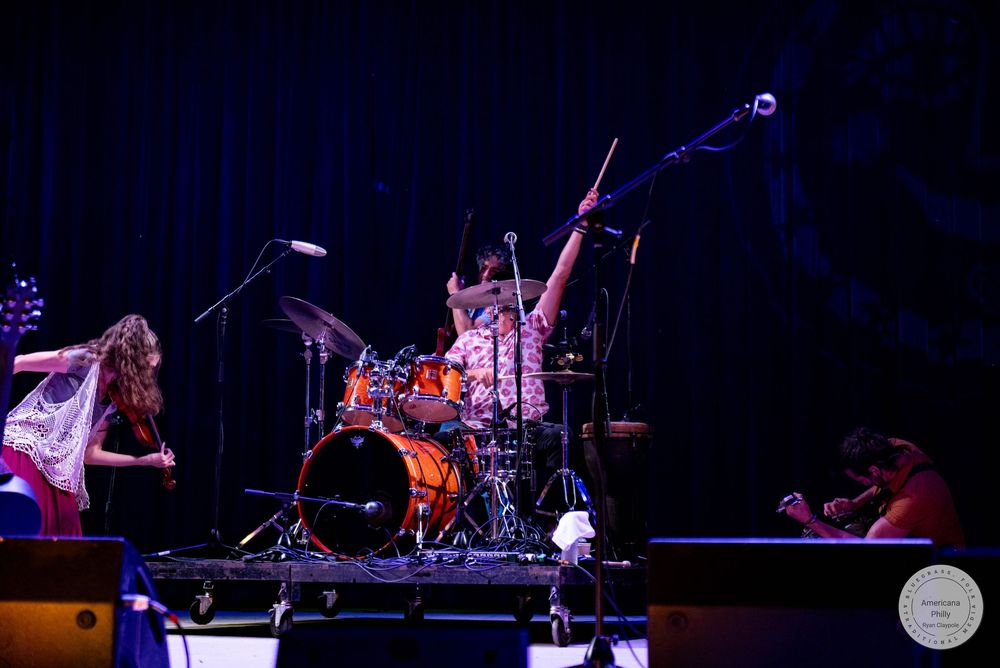 Band performing live on stage with drummer playing orange kit and guitarist under blue stage lighting.