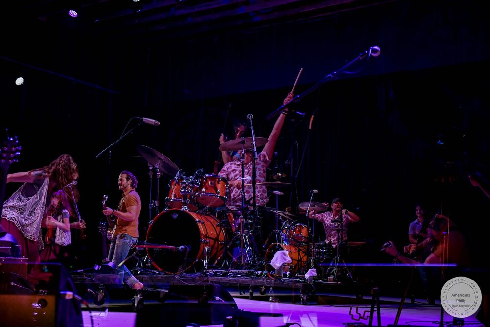 Live band performing on stage with dramatic purple lighting, drummer and guitarist visible in energetic concert setting.