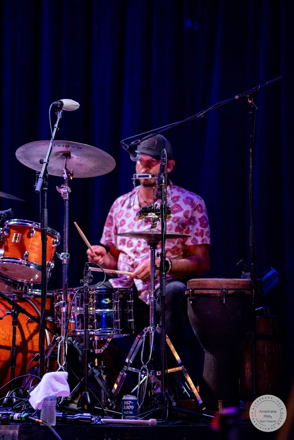 Male drummer in floral shirt playing drum kit on stage with cymbals and bongo under blue lights.