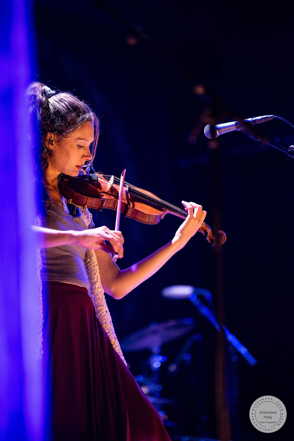 Young woman playing violin on stage under blue and purple stage lights wearing a red skirt at a live concert.
