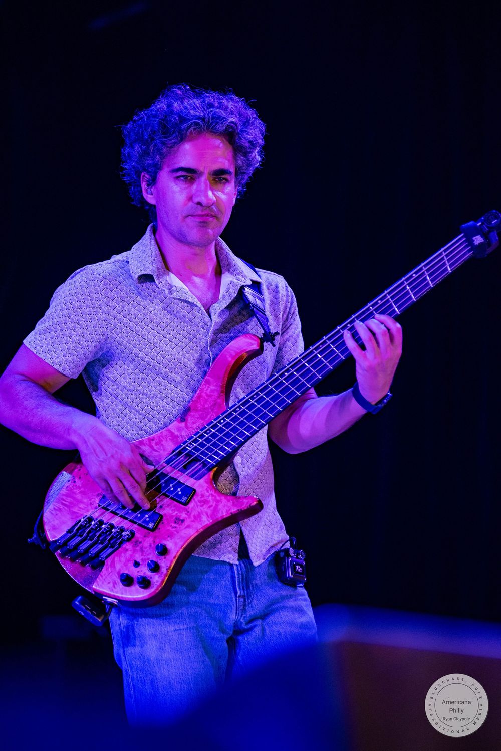 Curly-haired male bassist playing a red five-string electric bass guitar on a purple-lit stage.