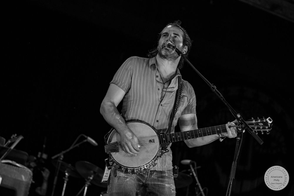 Man playing banjo and singing into microphone on stage in black and white concert photo.