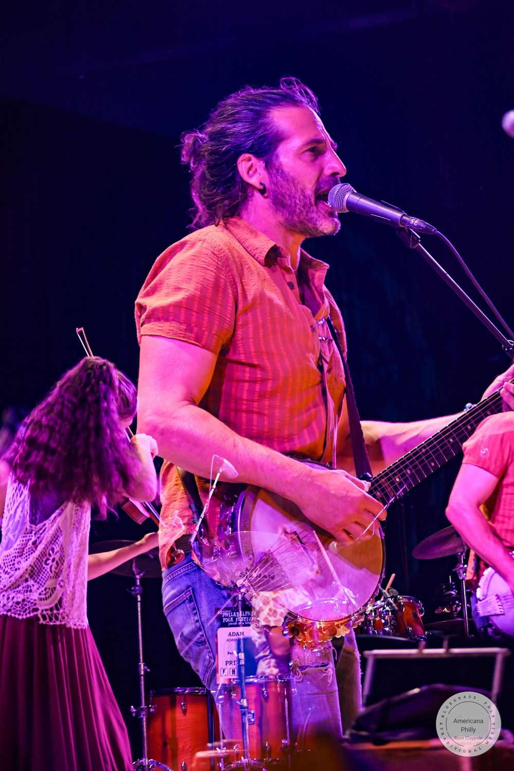 Bearded male guitarist singing into microphone on stage, wearing orange shirt, performing live concert under purple stage lighting.
