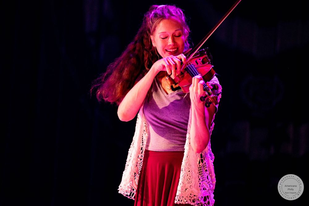 Young woman playing violin on stage under pink stage lighting, wearing a white lace vest and maroon skirt.