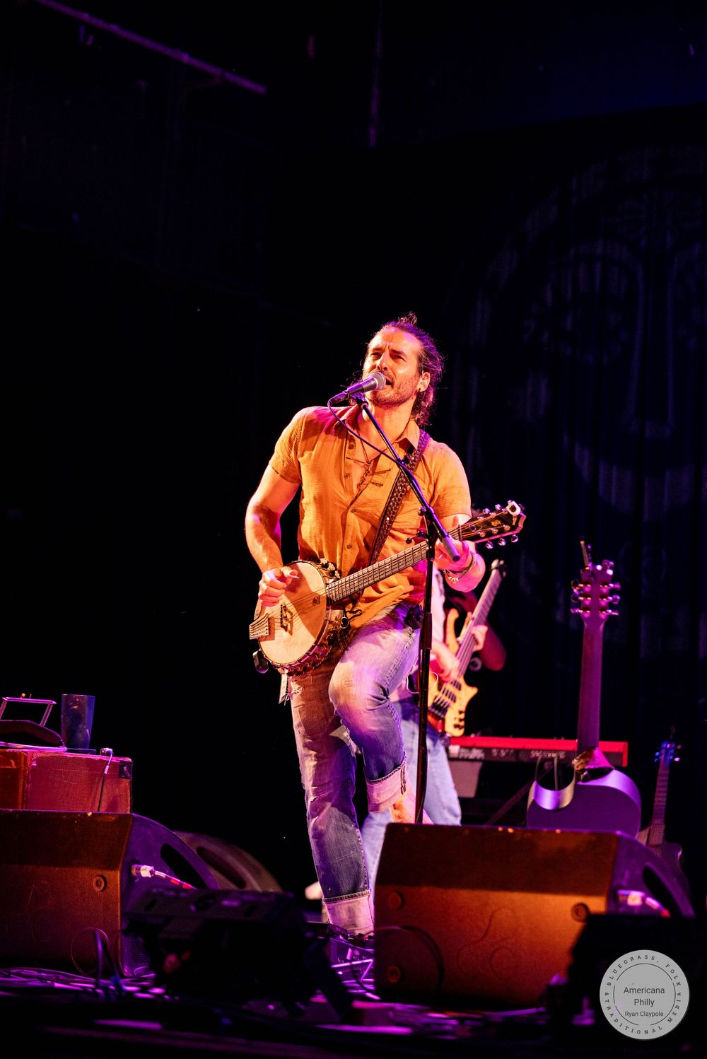 A male musician performs on stage playing a banjo guitar, wearing a yellow shirt and jeans under colorful stage lights.