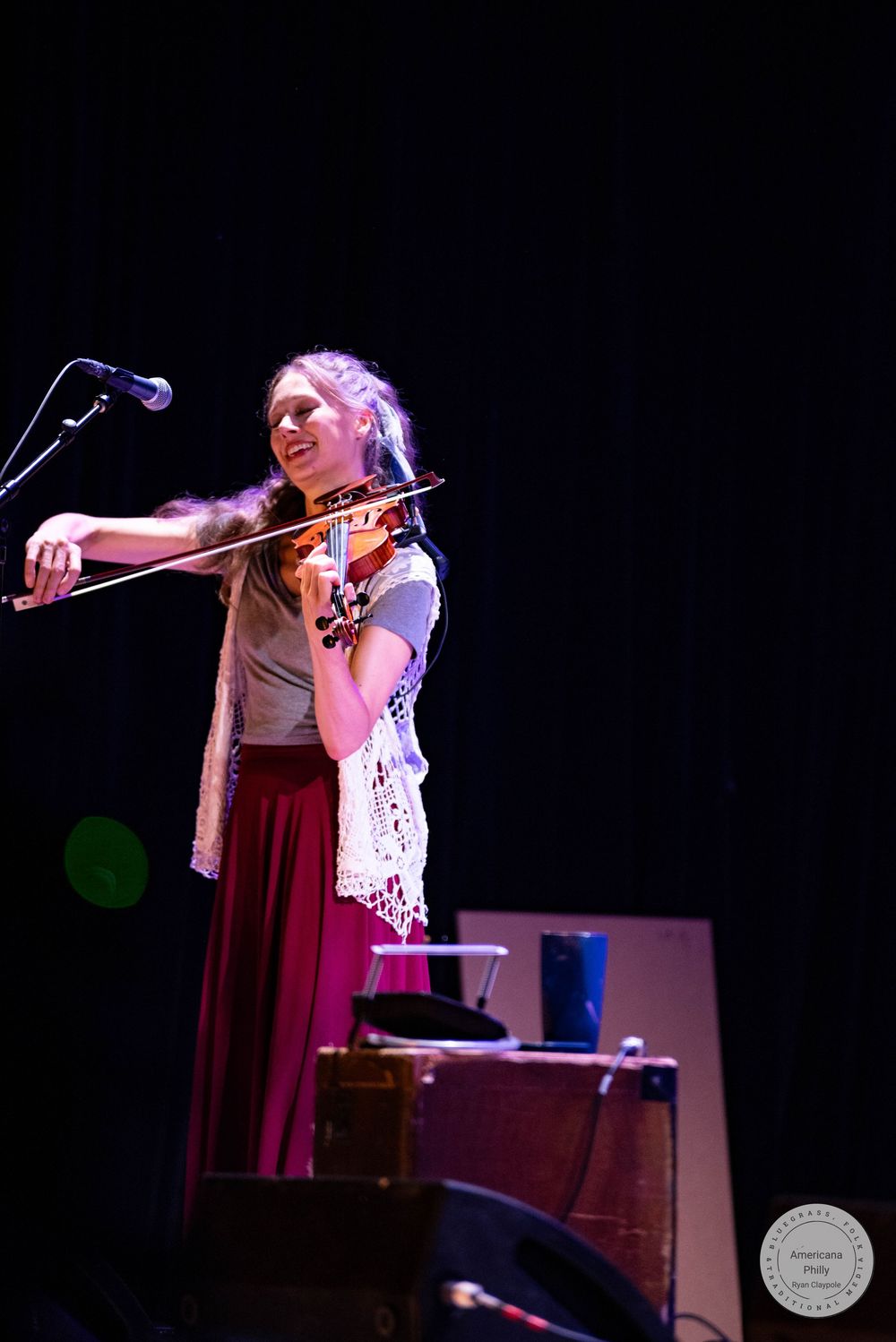 Young woman playing violin on stage, smiling, wearing a white lace vest and burgundy skirt under concert lighting.