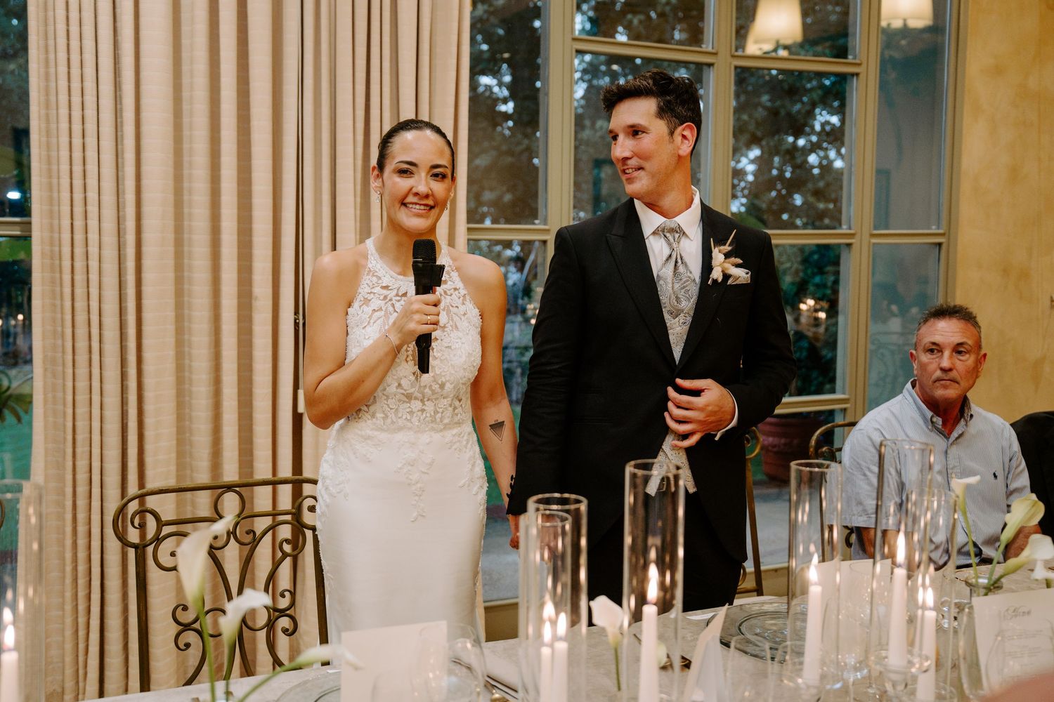 Bride speaks into microphone beside groom at elegant wedding reception dinner table with candles.