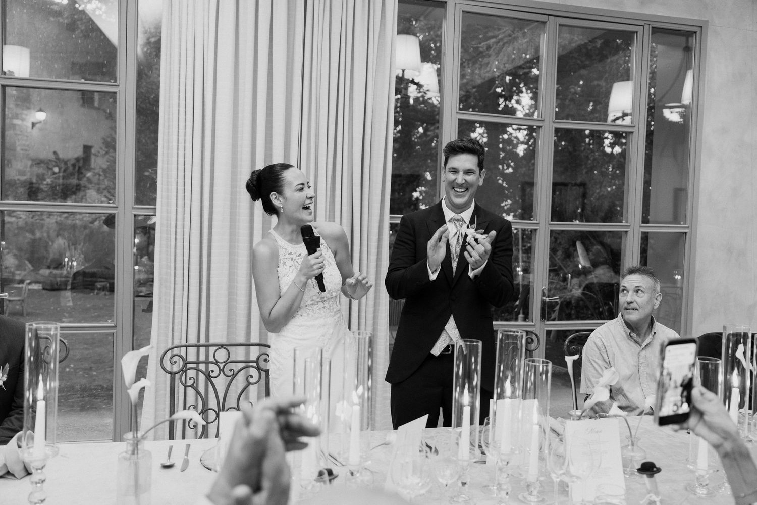 Bride speaks into microphone while groom applauds during elegant wedding reception dinner.