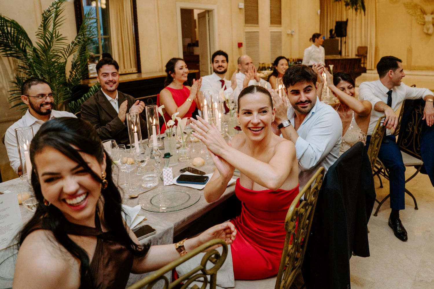 Wedding guests in formal attire smile and wave at camera during festive reception dinner celebration.
