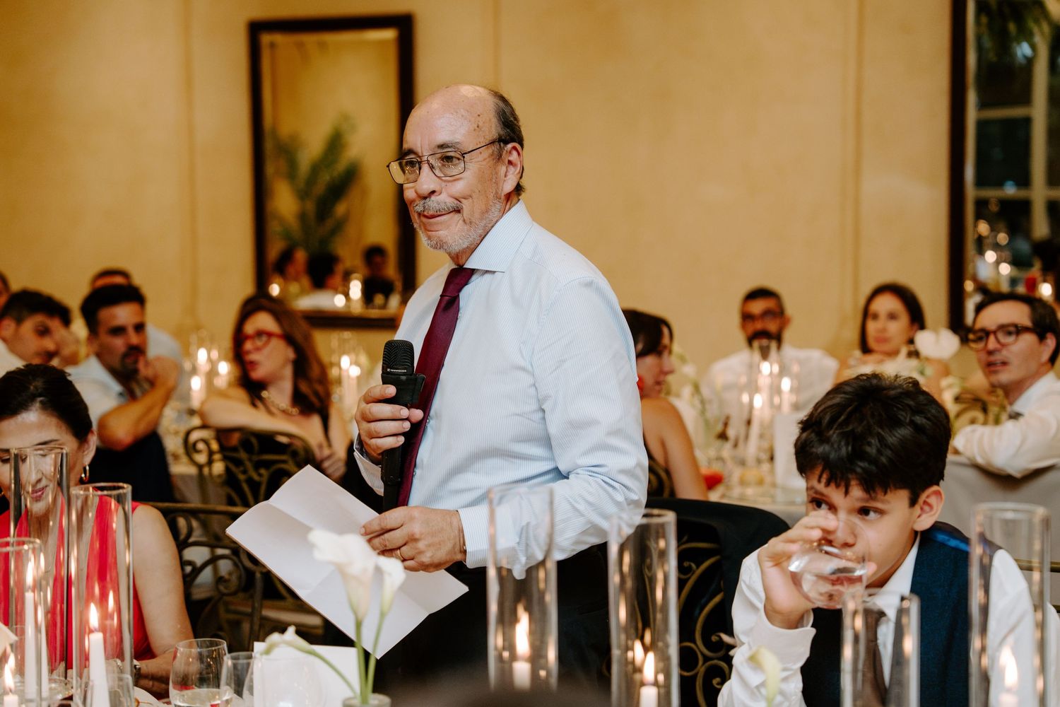 Man delivers heartfelt wedding speech holding notes while guests listen at candlelit reception dinner.