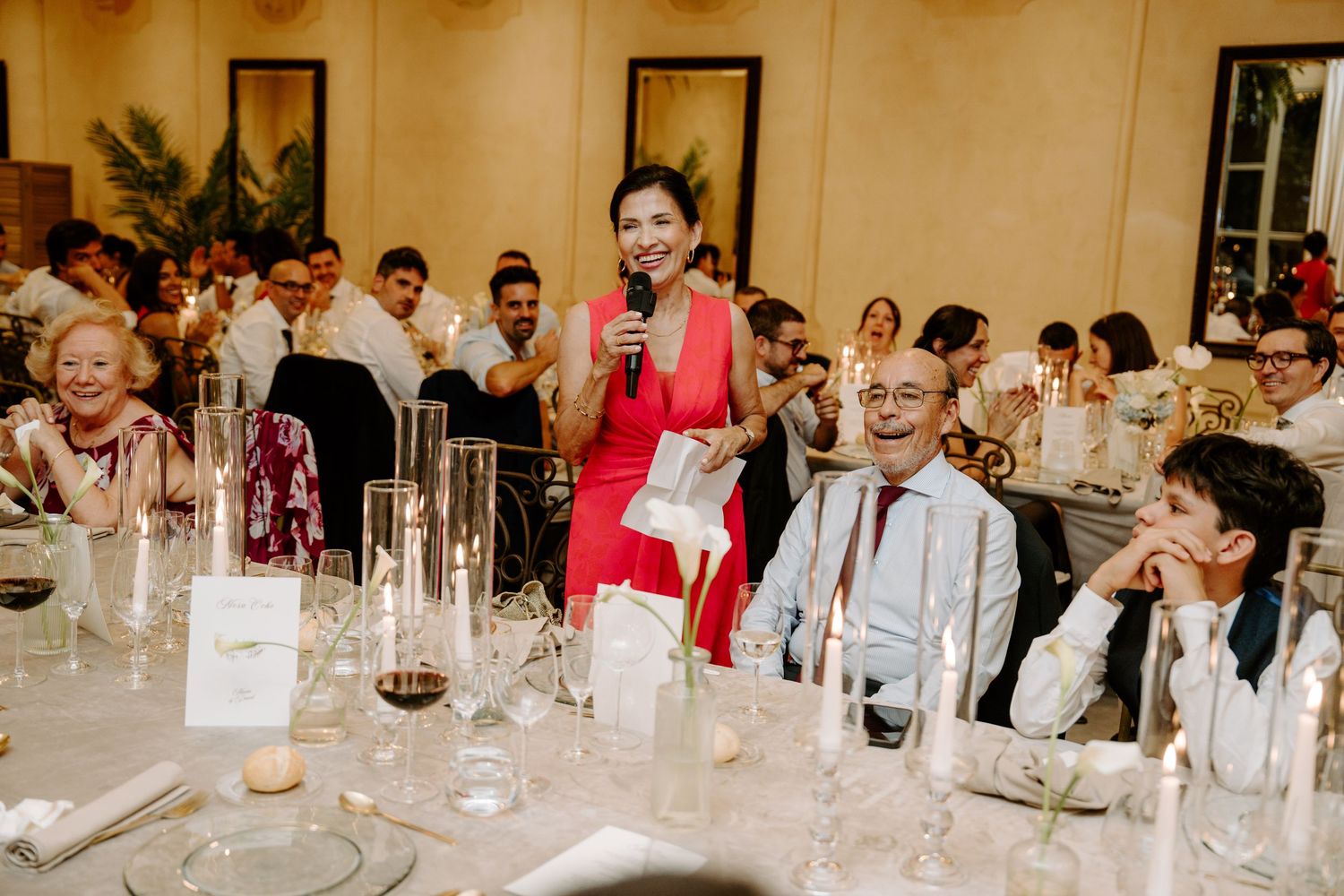 Woman in red dress giving a heartfelt wedding speech to guests seated at elegantly decorated reception tables.